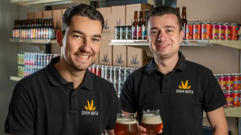 De oprichters Koen & Koen van GeheimBiertje in hun magazijn. Een portretfoto van Koen & Koen die proosten met een bierglas in hun hand en bedrijfskleding aan.